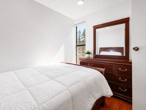 Un dormitorio con cama, cómoda con cajones, espejo y ventana con vista a árboles.