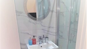 A bathroom with a glass-enclosed shower, a marble-tiled wall, a round mirror, and a white sink with two bottles of soap.