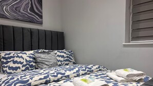 A neatly made bed with a blue and white patterned bedspread and pillows, a dark headboard, and a window with blinds.