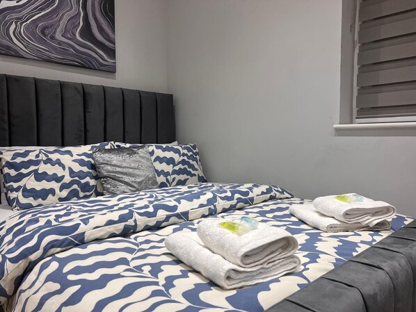 Una cama bien hecha con una colcha y almohadas de diseño azul y blanco, un cabecero oscuro y una ventana con persianas.