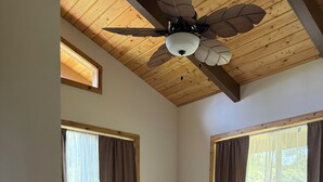 Une chambre avec un plafond en bois, un ventilateur de plafond, un lit recouvert d’une courtepointe à motifs et deux fenêtres avec des rideaux.