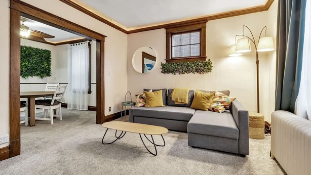 A cozy living room with a grey sectional sofa, a small round coffee table, and a dining area with a wooden table and chairs. - *Close to the Falls*Hot Tub*Easy Checkout*Parking* (Niagara Falls)