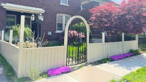 A two-story brick house with a white picket fence, a gated driveway, and a well-maintained front yard with purple flowers. - *Close to the Falls*Hot Tub*Easy Checkout*Parking* (Niagara Falls)