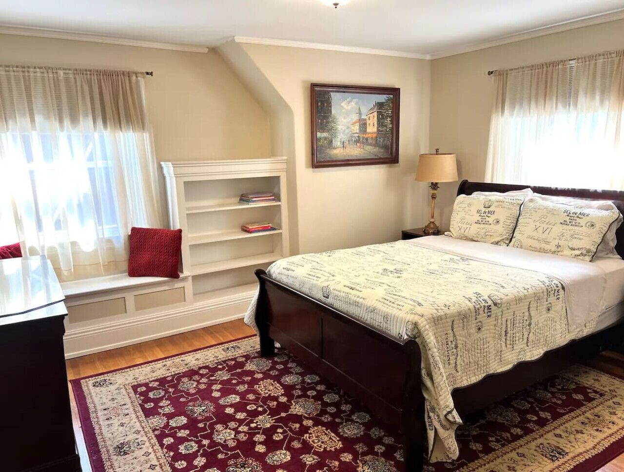 Une chambre avec un grand lit, un coin lecture près de la fenêtre avec des livres, un tableau au mur et un tapis rouge.