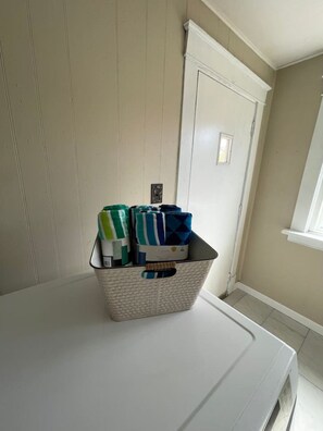 Una cesta blanca de ropa con prendas dobladas sobre un electrodoméstico blanco, probablemente una lavadora, en una habitación con paredes color beige y una puerta blanca.
