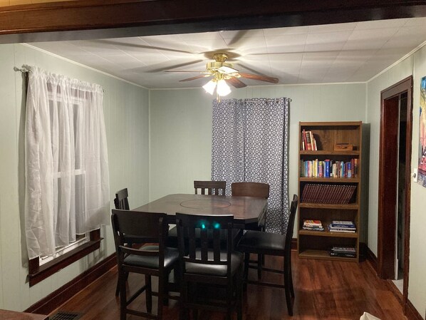 Une salle à manger avec une table et des chaises en bois, un ventilateur de plafond, une bibliothèque et une fenêtre ornée de rideaux légers.