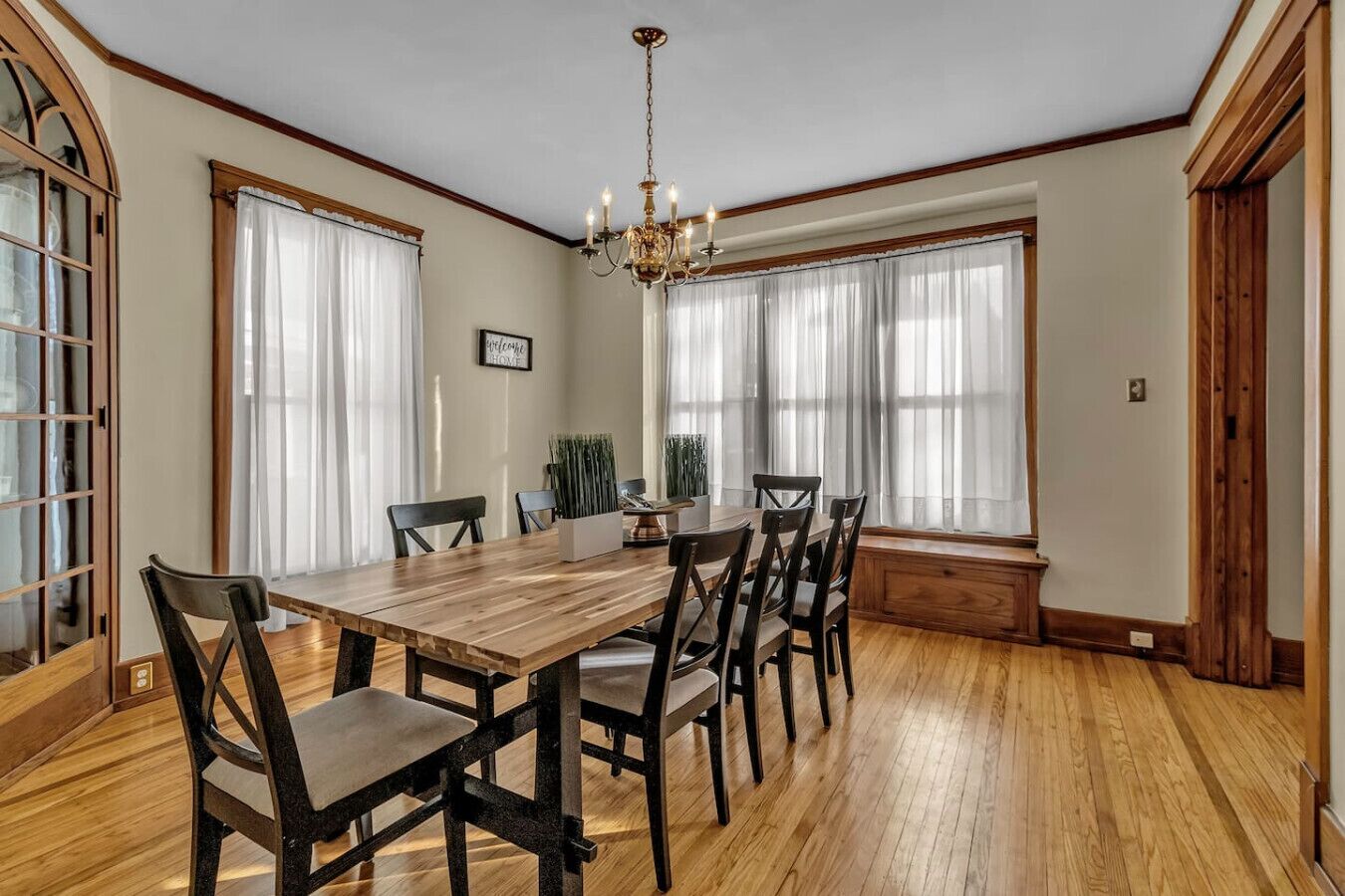A dining room with a wooden table and chairs, large windows with sheer curtains, and a chandelier.