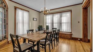 A dining room with a wooden table and chairs, large windows with sheer curtains, and a chandelier.