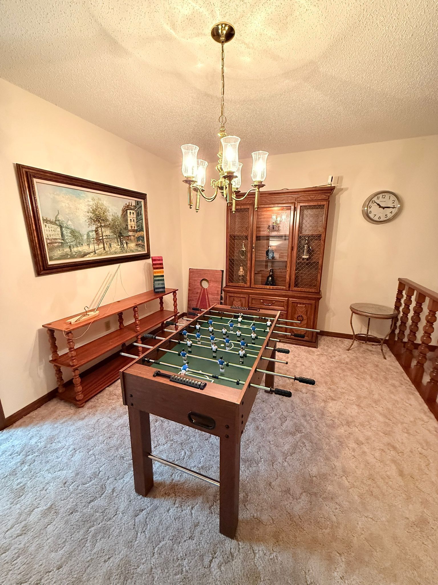 A room with a foosball table, a wooden bench, a chandelier, a clock, and a framed painting on the wall.