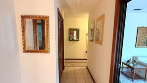 Un pasillo con suelo de baldosas, un espejo enmarcado, un ventanal pequeño y una puerta que da a una habitación con sofá y mesa.