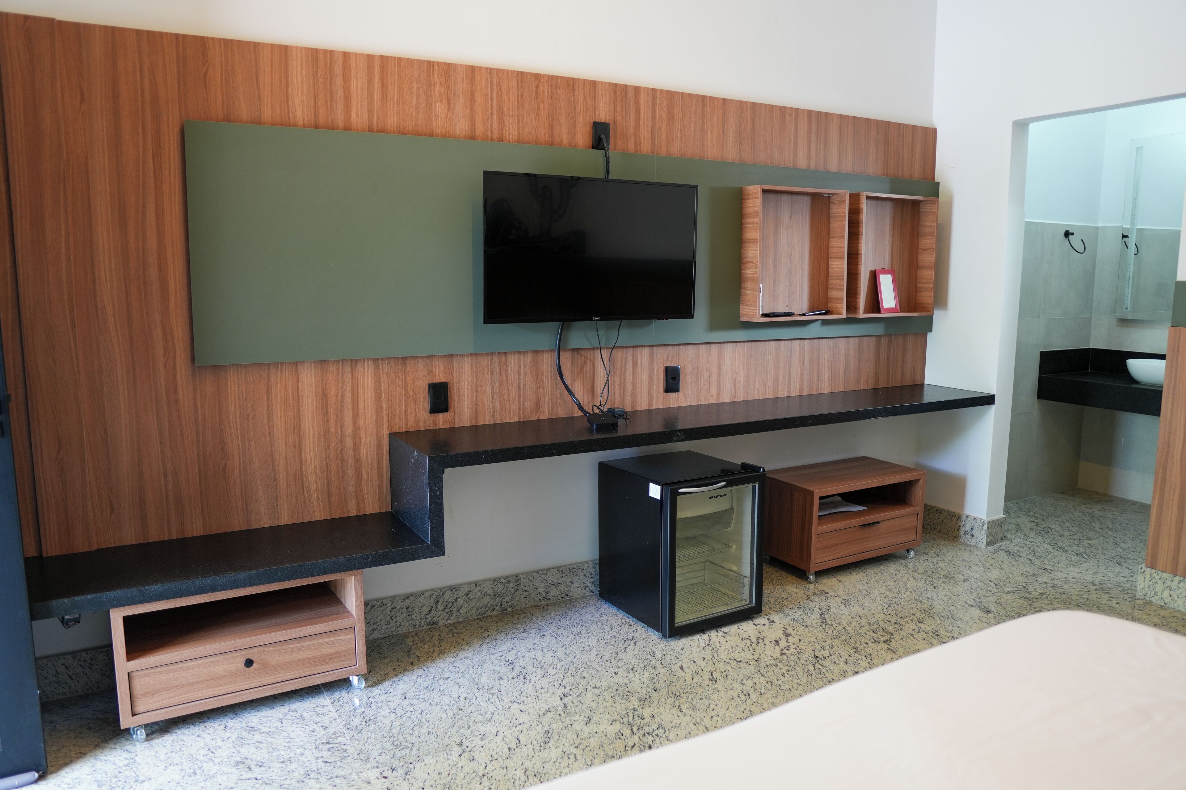 Een moderne hotelkamer met een houten accentmuur, een flatscreen televisie bevestigd boven een zwart aanrecht, een kleine koelkast en een houten kastje met spiegel.