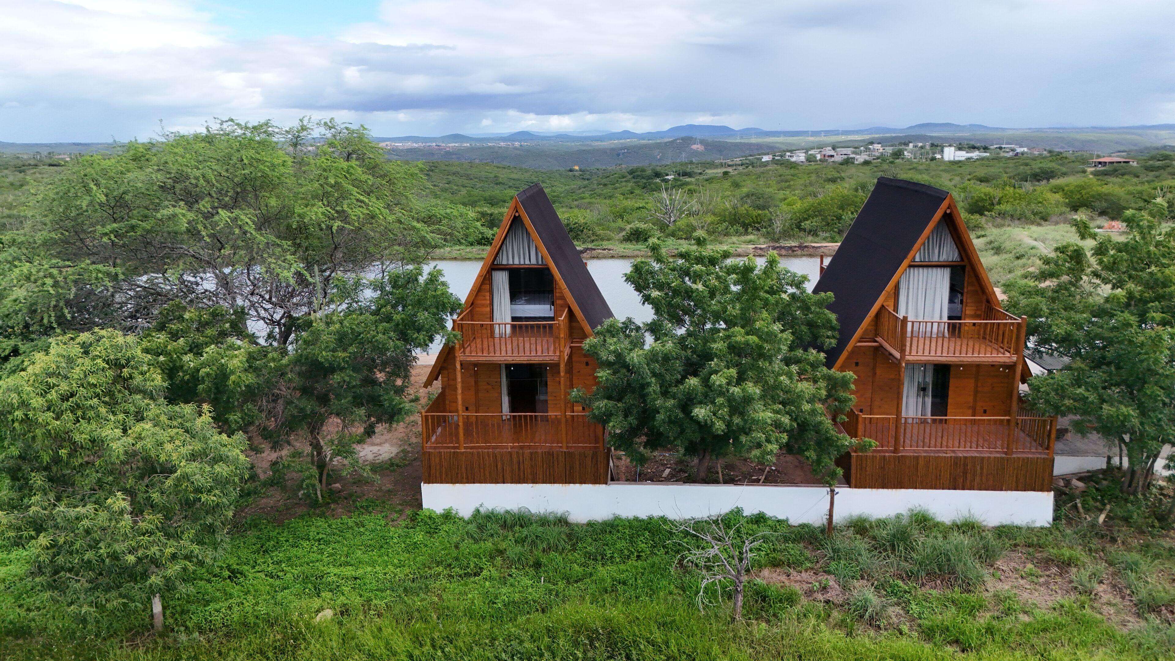 Duas cabanas de madeira com varandas, rodeadas por vegetação exuberante e um lago, com uma paisagem montanhosa ao fundo.