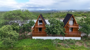 Duas cabanas de madeira com varandas, rodeadas por vegetação exuberante e um lago, com uma paisagem montanhosa ao fundo.