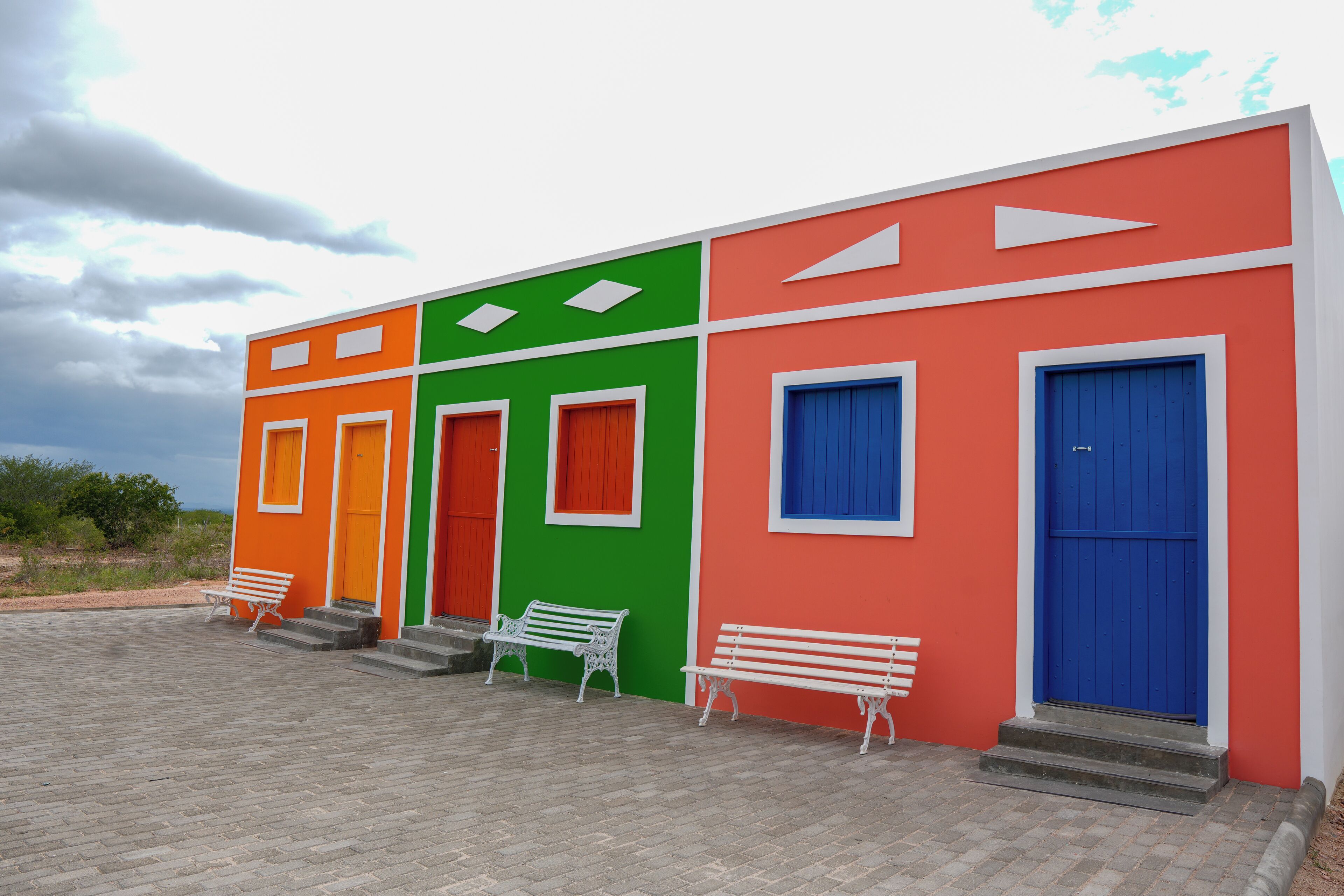 A building with four doors, each painted a different color: orange, green, red, and blue. There are steps leading up to each door, and two white benches are placed in front of the doors.