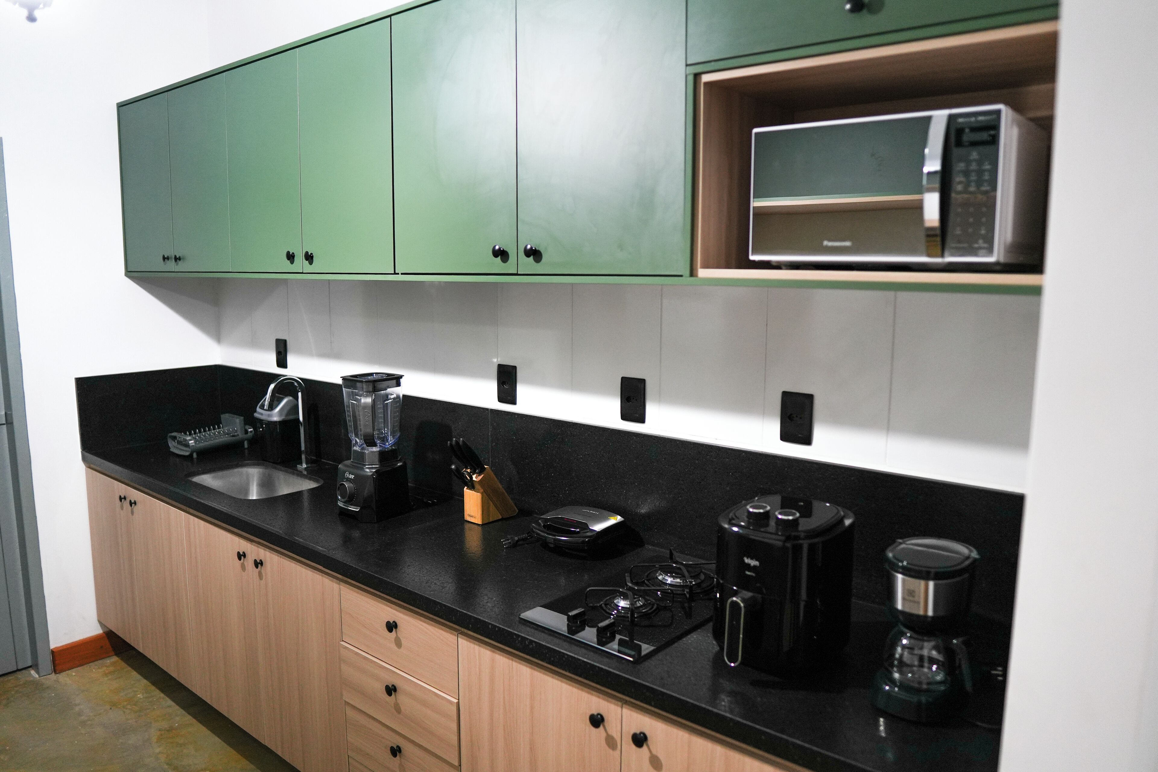 A kitchen with green cabinets, a black countertop, and various appliances including a microwave, coffee maker, and blender.