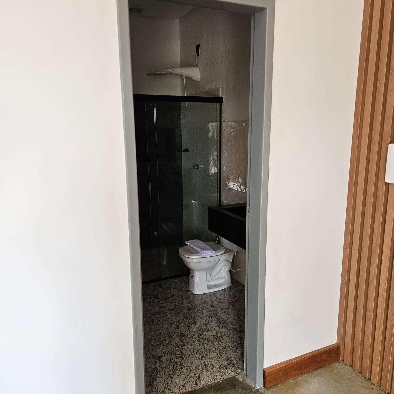 Uma porta estreita dá acesso a um banheiro com vaso sanitário, box de vidro e pia branca.