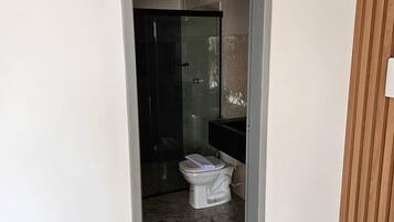 Uma porta estreita dá acesso a um banheiro com vaso sanitário, box de vidro e pia branca.
