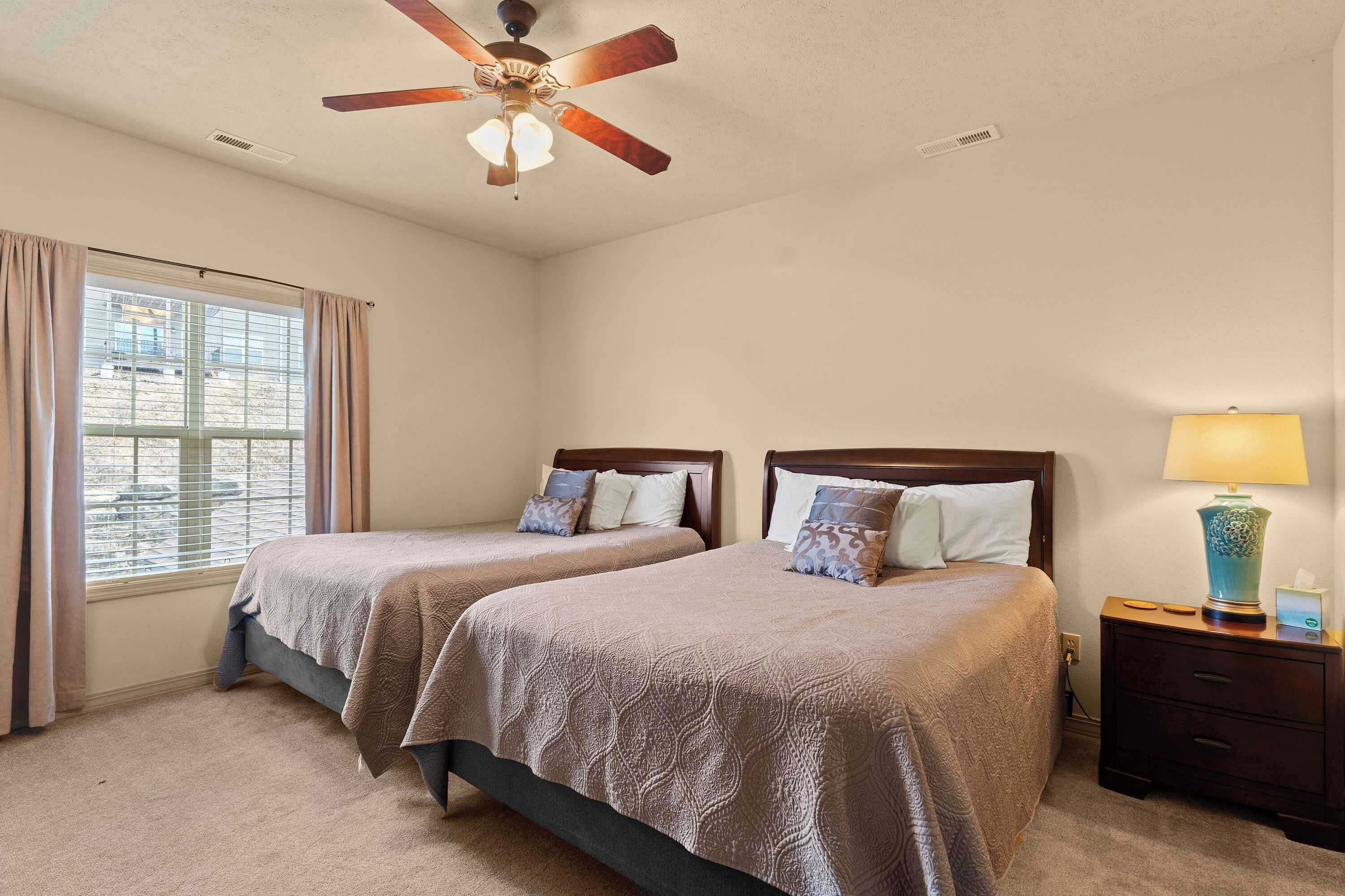 A hotel room with two beds, a ceiling fan, a window with curtains, a bedside table with a lamp, and a view of the outside.