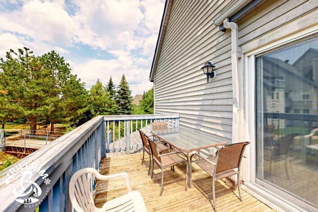 A wooden deck with a glass table and chairs, a sliding glass door, and a view of trees and a cloudy sky.
