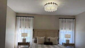 A bedroom with a large bed, two bedside tables with lamps, a chandelier, and sheer curtains.