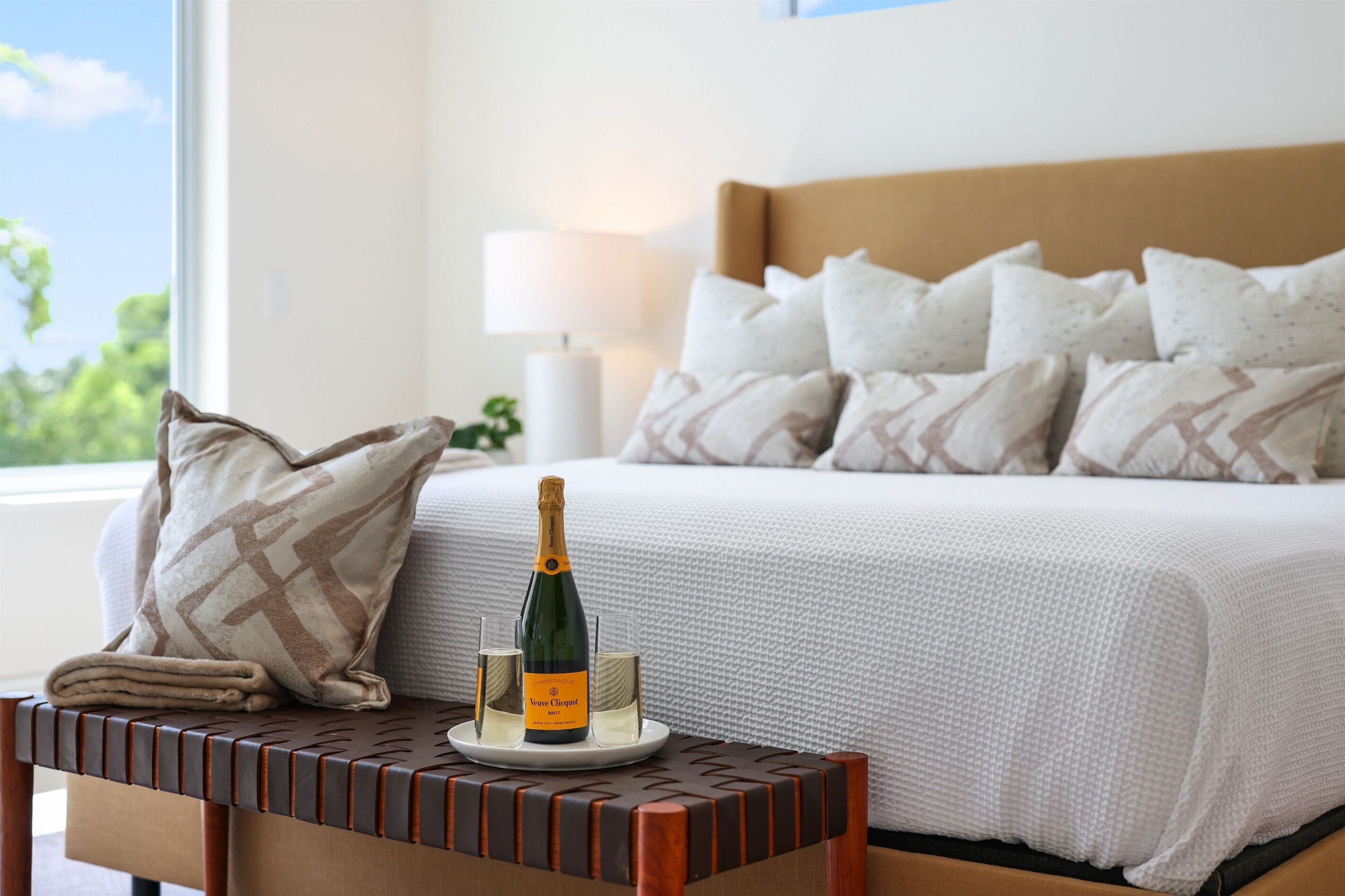 A neatly made bed with a white textured bedspread and multiple pillows, a bottle of champagne on a small table beside the bed, and a lamp on a bedside table.