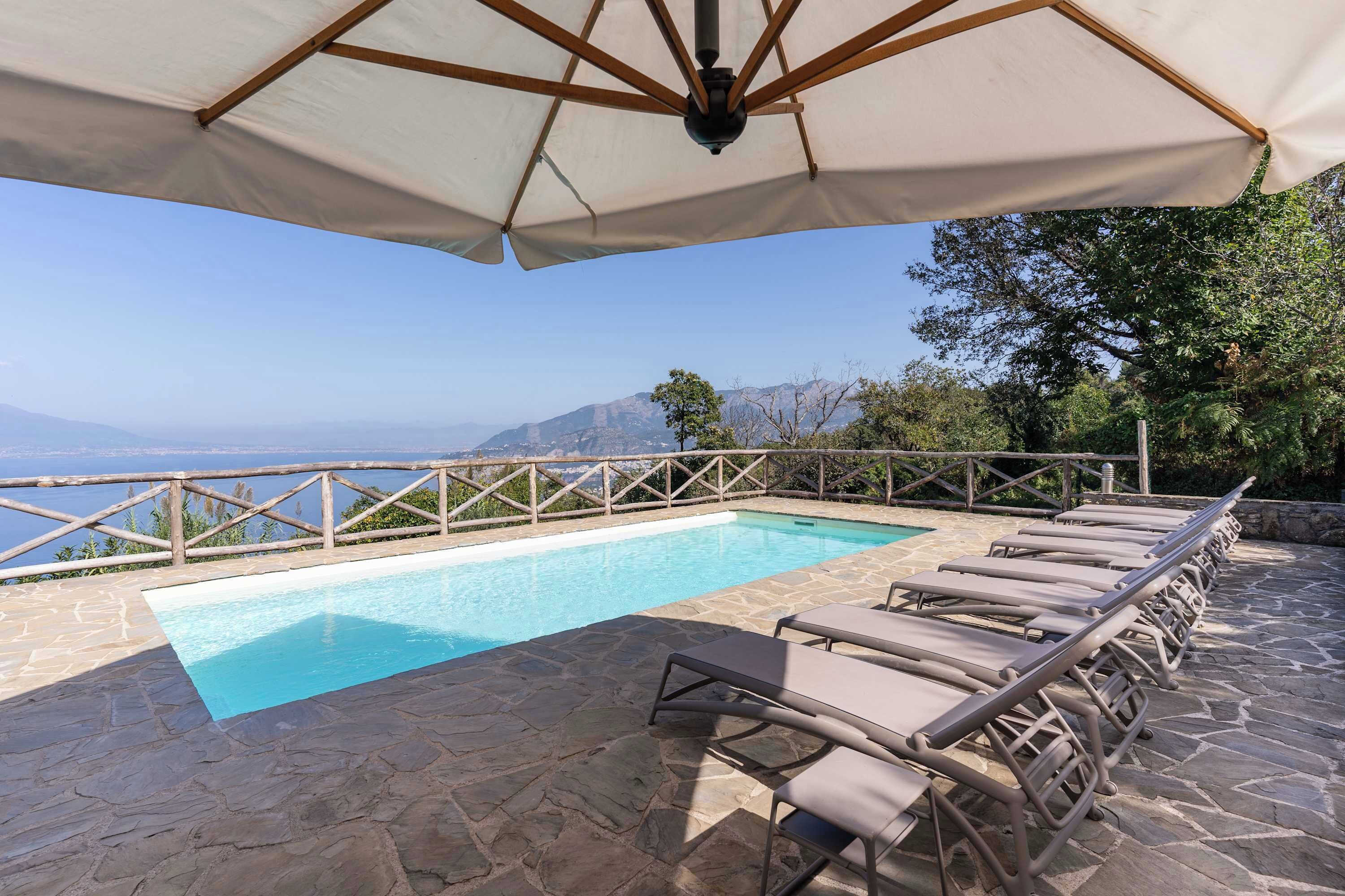 Piscina com espreguiçadeiras e guarda-sol, cercada por pavimento de pedra e cerca de madeira, com vista para uma paisagem deslumbrante.