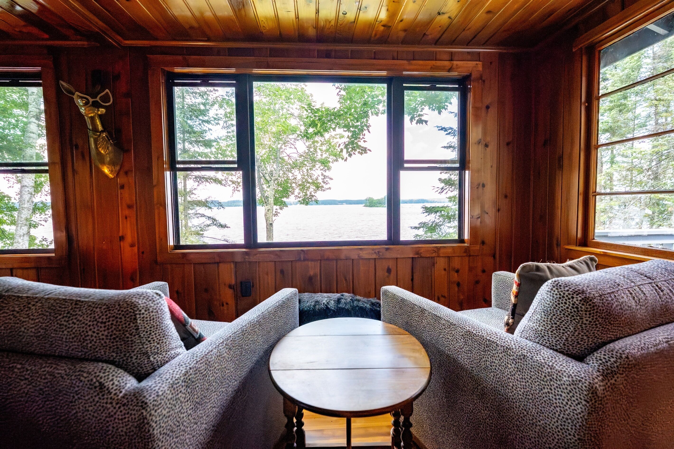 A cozy cabin interior with wooden walls, a large window offering a view of a lake, and a round table in the center.