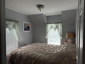 Un dormitorio con una cama grande, dos ventanas con cortinas, una lámpara de techo y un cuadro en la pared.