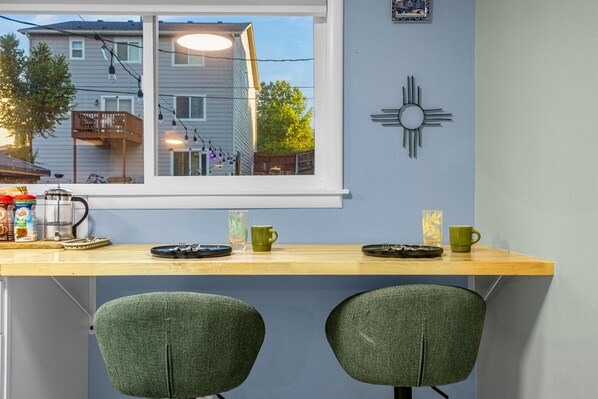 A kitchen with a wooden countertop, two green chairs, a window with a view of a house, and a wall-mounted decorative item.