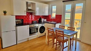 A compact kitchen with white cabinets, a red-tiled backsplash, a built-in oven, a washing machine, and a dining area with a table and chairs.