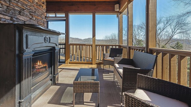 Una terraza de madera con un muro de piedra, una chimenea, muebles de mimbre y grandes ventanales que ofrecen vistas a las colinas.