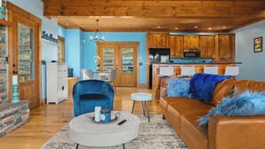 Amplia sala de estar con un sillón azul, un sofá de cuero marrón y una mesa de centro redonda. Se observa una cocina con armarios de madera y un microondas al fondo.