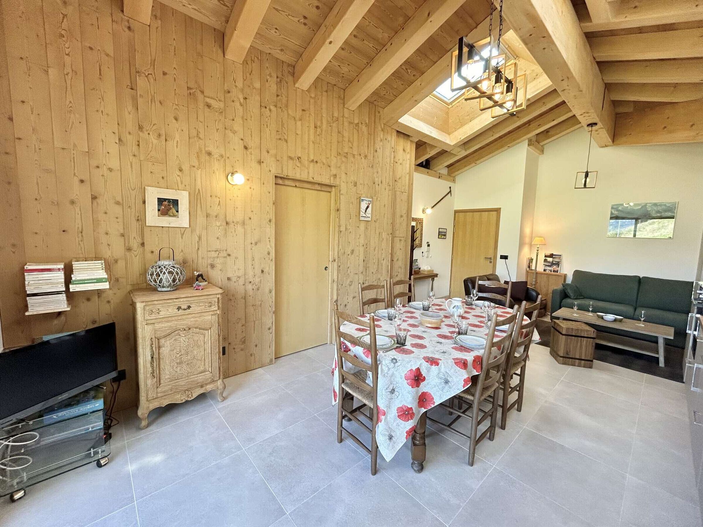 Une pièce chaleureuse avec un plafond en bois, une table à manger dressée pour quatre personnes, un canapé et un téléviseur.