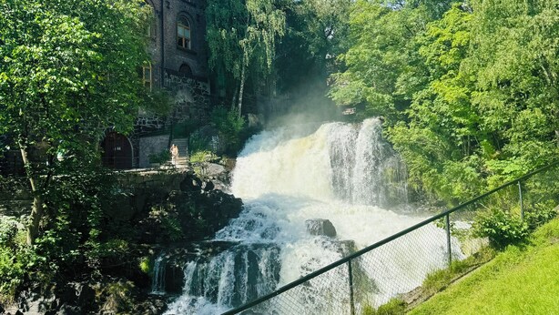 Een historisch gebouw met meerdere ramen en een bakstenen gevel, met uitzicht op een waterval en weelderige begroeiing.