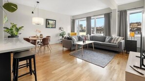 Een moderne woonkamer met een bank, eettafel en stoelen. Er is een groot raam met gordijnen, een plafondlamp en een plant aan de linkerkant.