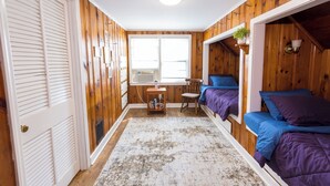 Ein Zimmer mit Holzwänden, einem Fenster, einem Schreibtisch mit Stuhl und zwei Betten mit violetter Bettwäsche.