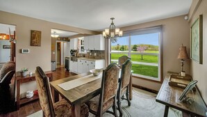 A dining area with a wooden table and chairs, a chandelier, and a view of a green landscape through large windows.