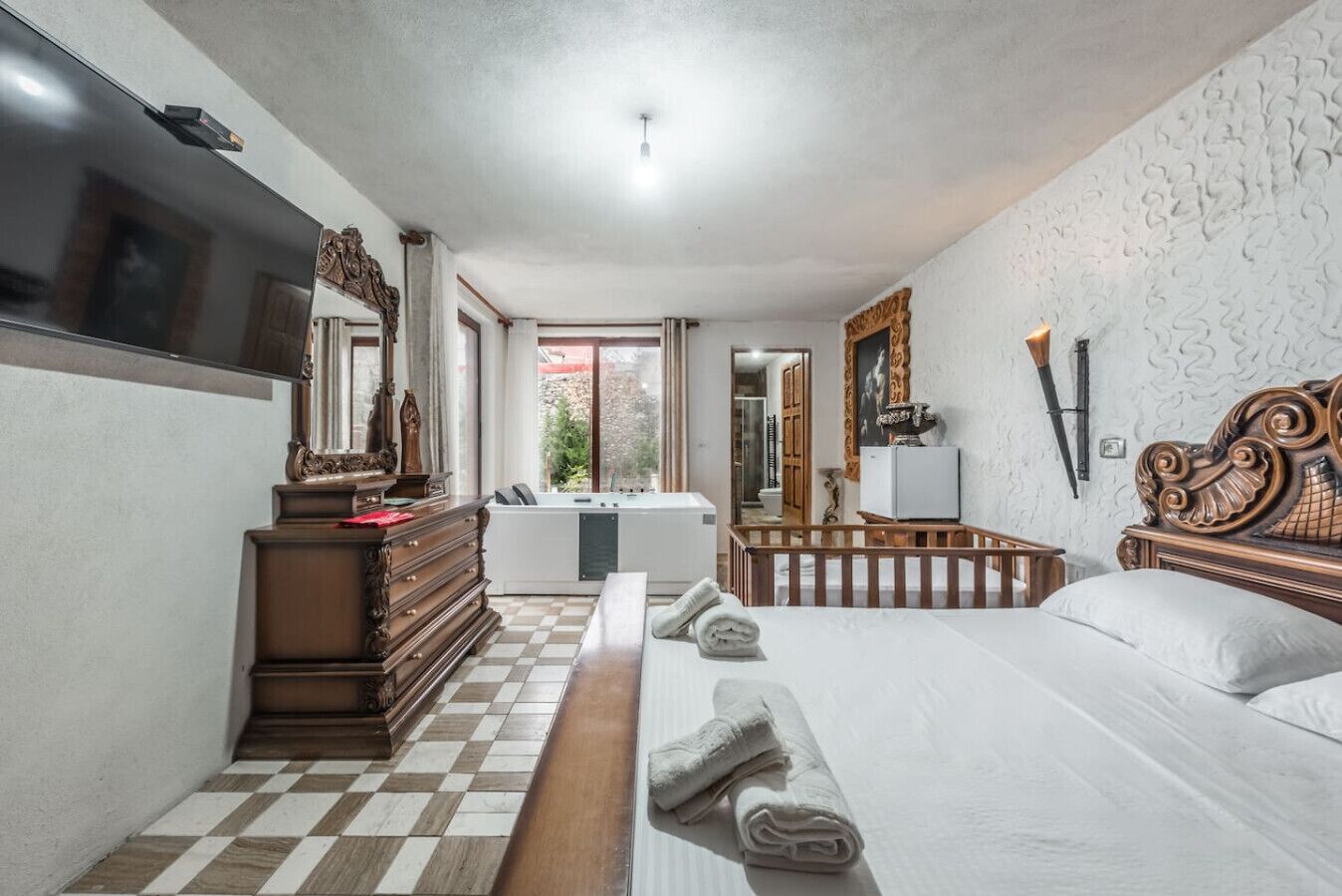 A hotel room with a checkered floor, a bed with neatly folded towels, a wooden dresser, a television, and a large mirror.