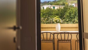 Vista desde una habitación con puerta, hacia un balcón con un jarrón blanco lleno de flores y dos sillas de mimbre.