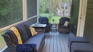 A covered outdoor seating area with a sofa, chairs, and ottomans, featuring a view of a garden and trees.