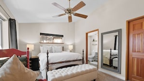 Quarto de hotel com cama, um banquinho, uma cadeira, ventoinha no teto, um espelho grande e uma porta de madeira.
