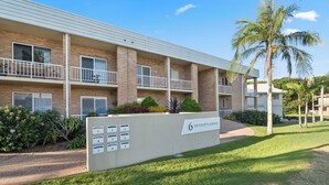 A multi-story apartment building with numbered mailboxes, a sign indicating '6 on Fourth Avenue', and a well-maintained lawn with palm trees. - Sunray – Beachside Bliss Sawtell, Beach, Village (Sawtell)