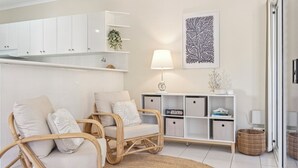 A modern living room with two wicker chairs, a white sofa, a woven rug, a white cabinet with shelves, a wall-mounted lamp, and a framed artwork. - Sunray – Beachside Bliss Sawtell, Beach, Village (Sawtell)