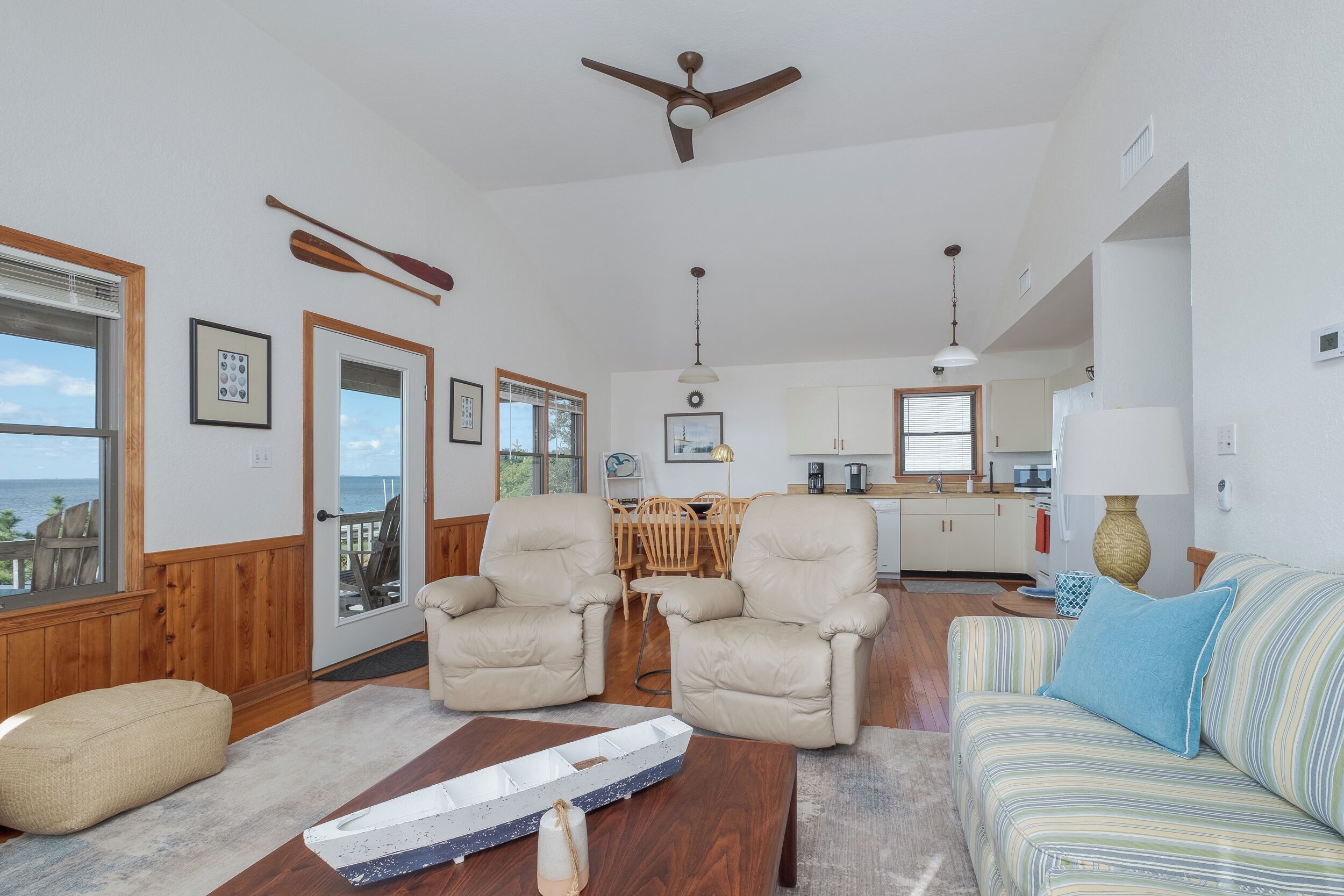 A spacious living room with a view of the ocean, a ceiling fan, a dining area, and a kitchen.