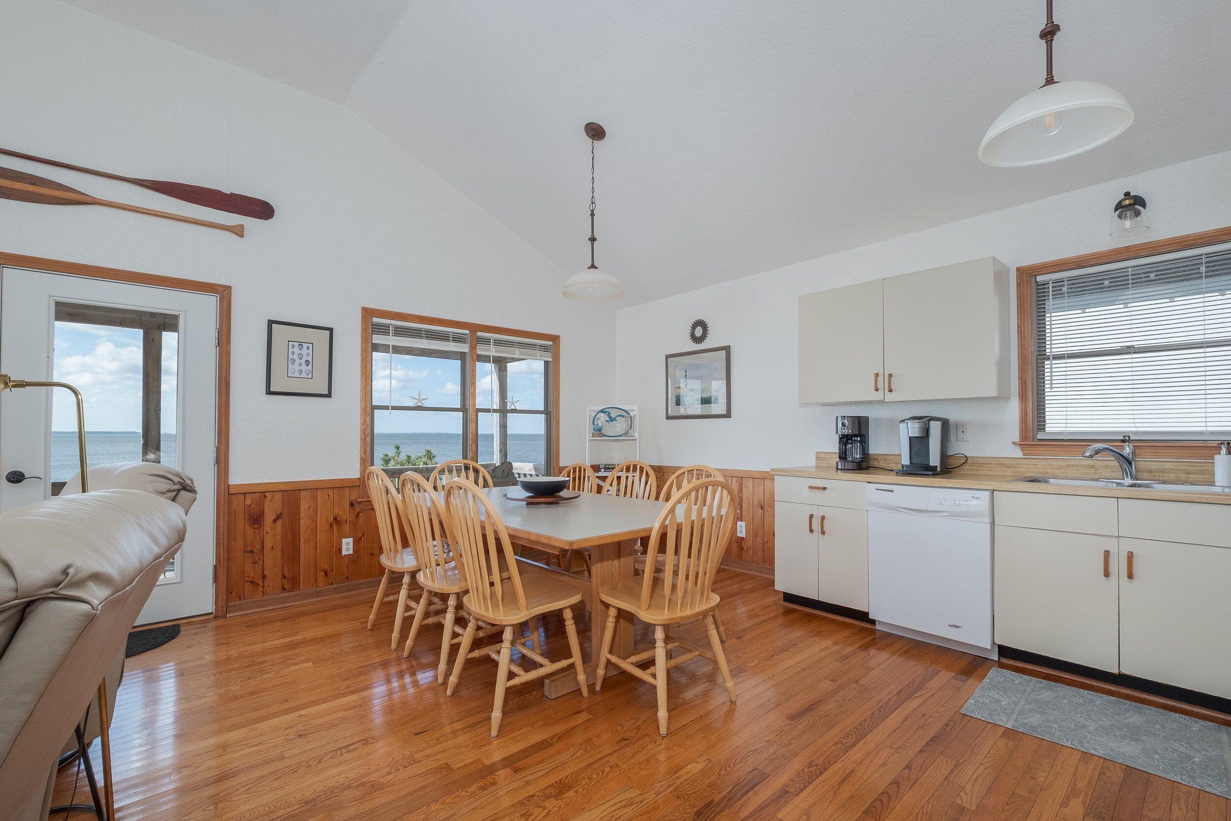Un comedor con mesa y sillas de madera, una cocina con armarios blancos y vistas al mar a través de amplios ventanales.