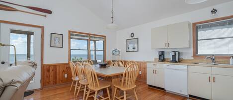Un comedor con mesa y sillas de madera, una cocina con armarios blancos y vistas al mar a través de amplios ventanales.