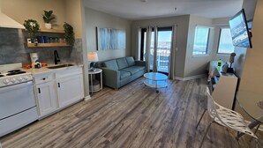 A modern kitchen with white cabinets, a built-in stove, and a small island with a sink. There is a grey sofa and a round glass table in the living area, with a balcony visible through the window. - Beachy 7th floor studio @ Myrtle Beach Resort (Myrtle Beach)