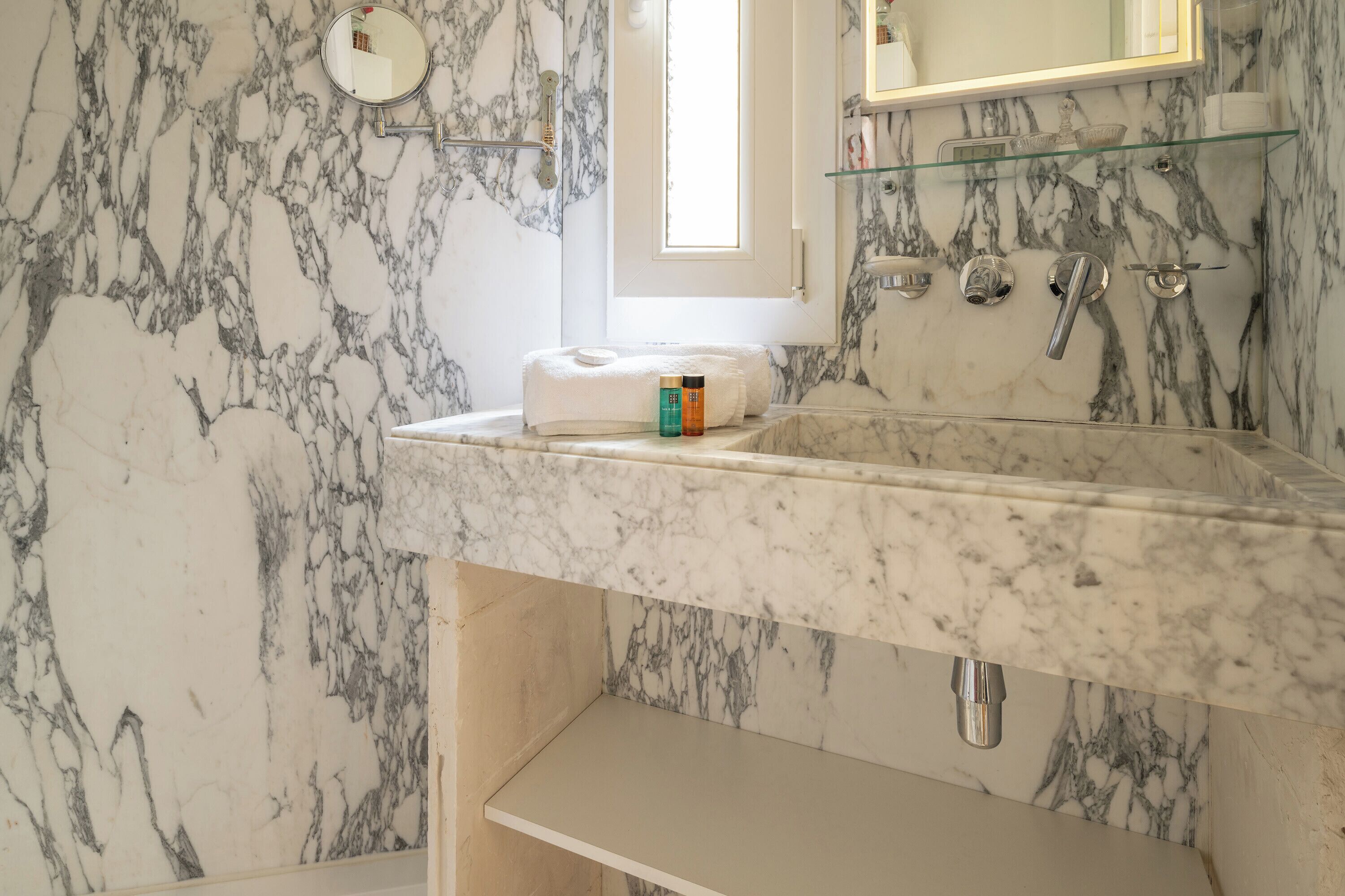 A bathroom with marble walls, a sink with a glass shelf, and a towel on the counter.