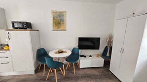 Un espace de vie compact comprenant un coin cuisine, un coin repas avec deux chaises turquoise et une petite table ronde, un téléviseur à écran plat fixé au mur et une grande armoire.