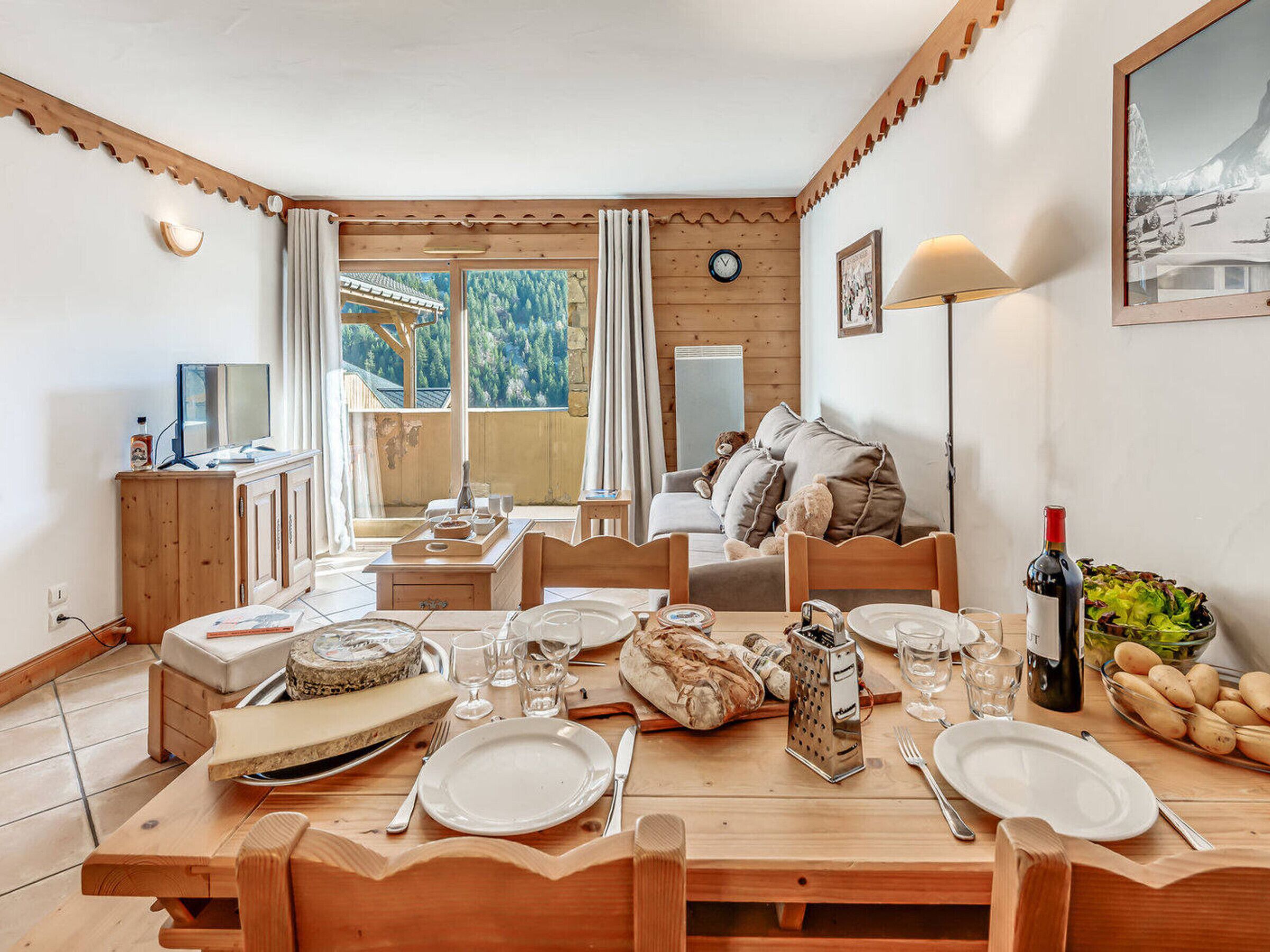 Un rincón comedor acogedor con una mesa de madera lista para una comida, un sofá con cojines, un reloj en la pared y un ventanal con vistas a árboles.